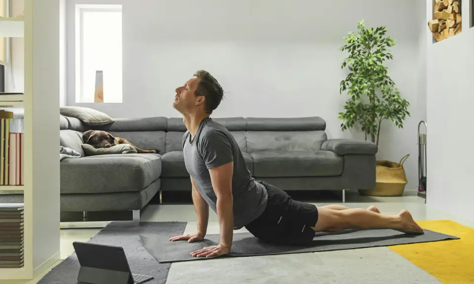 Man in cobra pose op yogamat, neemt regie over zijn leven. Hond slaapt op bank.