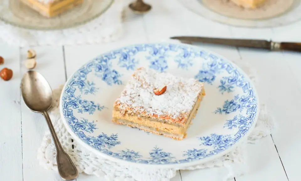 Un carré de gâteau russe, saupoudré de sucre glace et garni d'une noisette, sur une assiette ornée de bleu.
