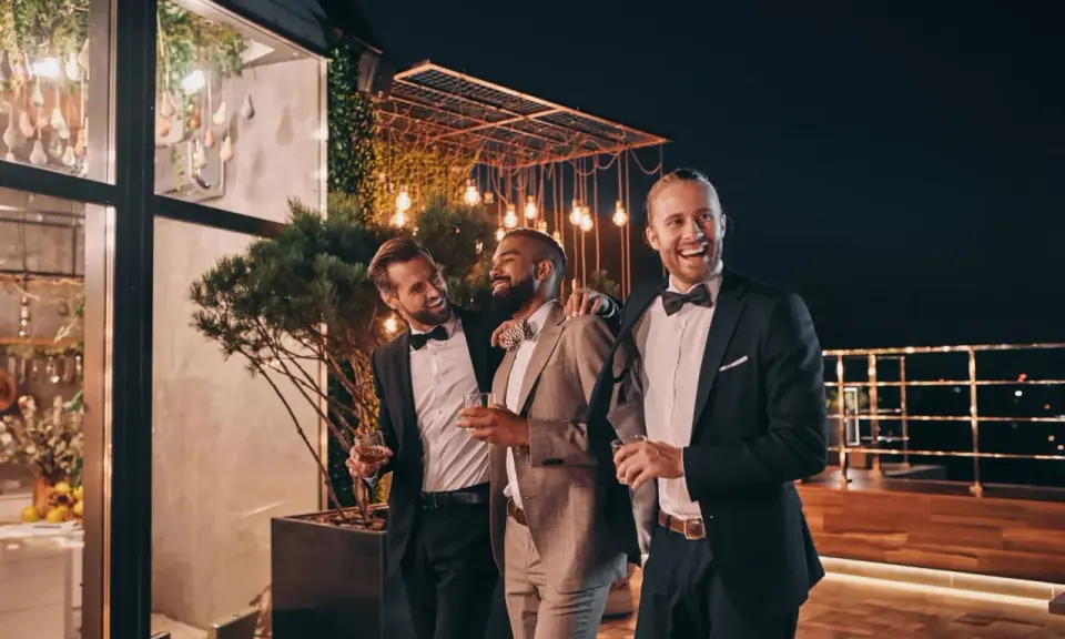 Trzech eleganckich panów, w tym drużba na weselu, śmieje się i trzyma drinki na tarasie nocą, wśród ozdobnych świateł.