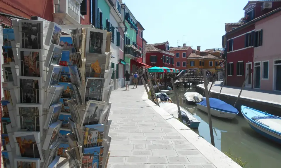 Cartoline di Murano, Burano e Torcello in vendita lungo un canale con case colorate e barche ormeggiate.