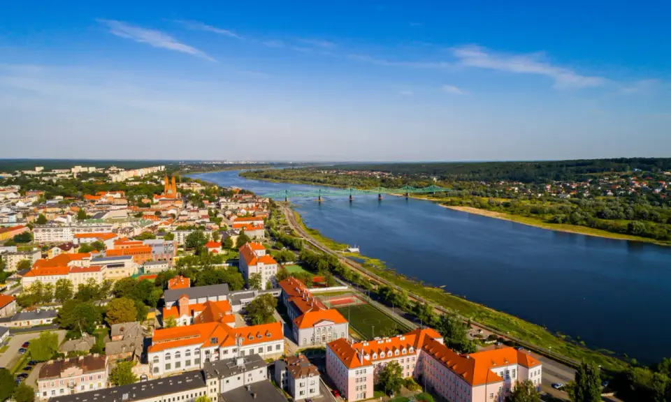 Widok z lotu ptaka na miasto nad Wisłą, z zielonym mostem i zabudowaniami w słoneczny dzień.