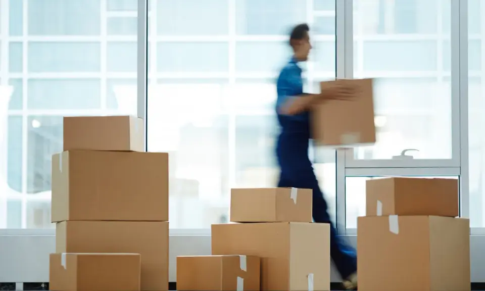 Un déménageur porte une boîte, entouré de cartons empilés. Une scène typique pour une facture déménagement.