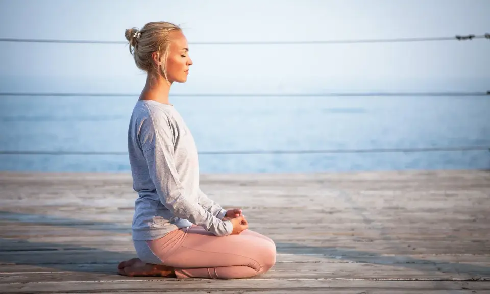 Kvinna med slutna ögon sitter i meditationsställning på en träbrygga vid havet, utövar progressiv avslappning.