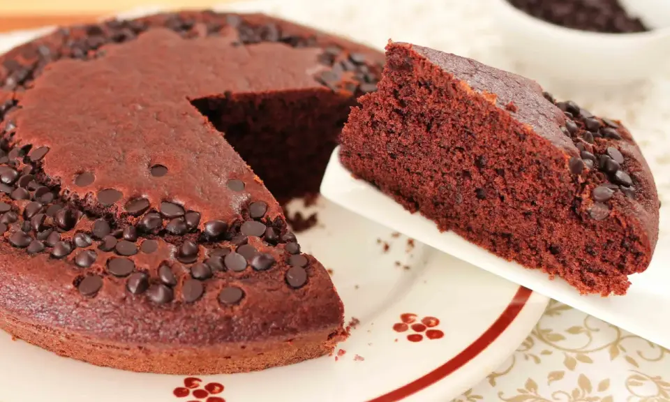 Una fetta di torta al cioccolato morbida, cosparsa di gocce di cioccolato, viene servita da una tortiera intera.