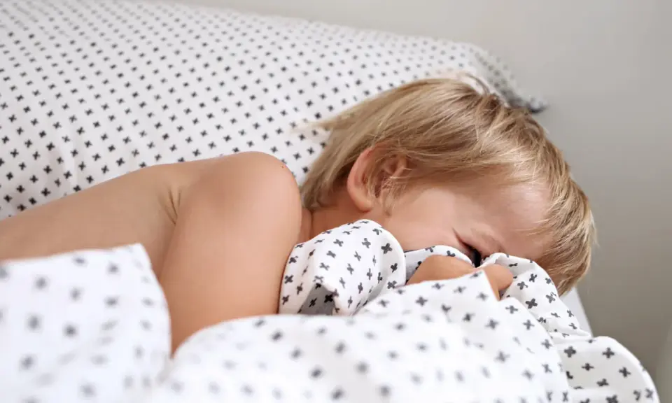 Niño rubio se esconde bajo la sábana. Aprende como hacer que un niño duerma solo con paciencia y rutinas.