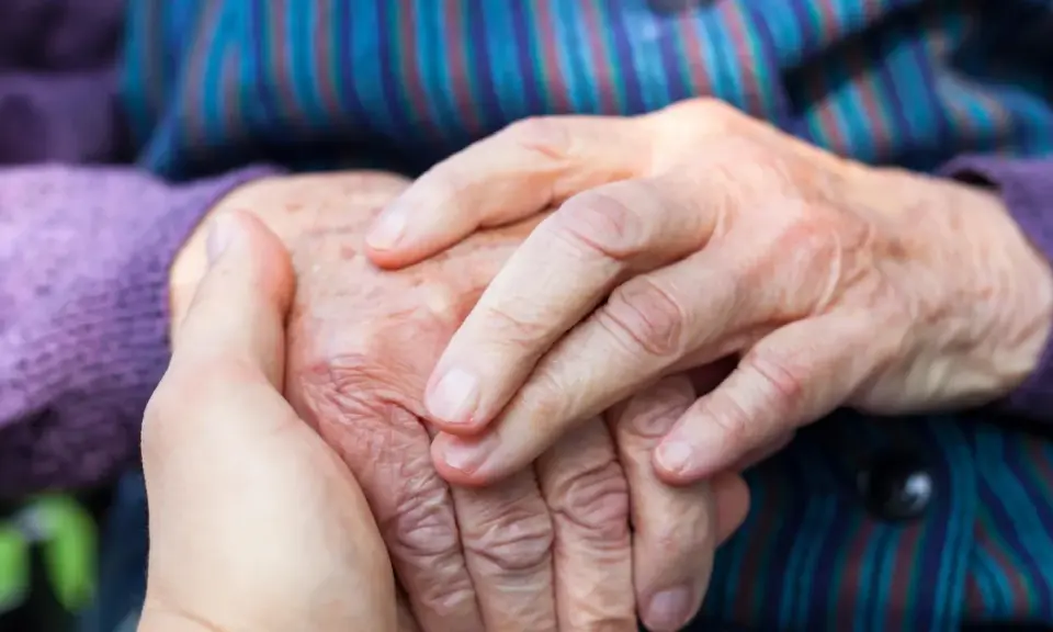 Dłonie młodszej osoby obejmują dłoń starszej, symbolizując wsparcie w obliczu choroby Alzheimera i jej przyczyn.