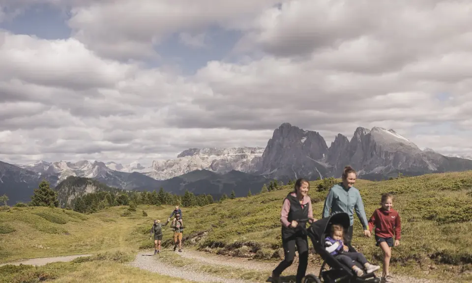 Famiglia in escursione sull'Alpe di Siusi, con bambini e passeggino, sullo sfondo le maestose vette dolomitiche.