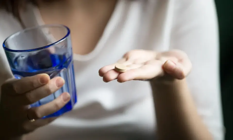 Une main tient un comprimé, peut-être un supplément de sels minéraux, à côté d'un verre d'eau bleue.