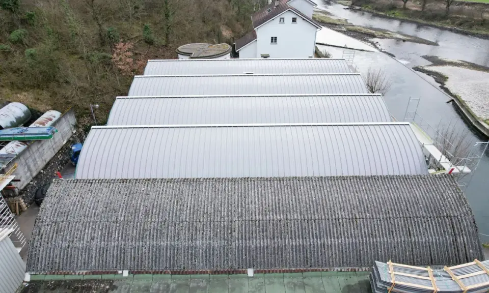 Reihen von Sheddach-Konstruktionen mit Metall- und Wellplatten. Ein Fluss und ein Gebäude sind im Hintergrund zu sehen.