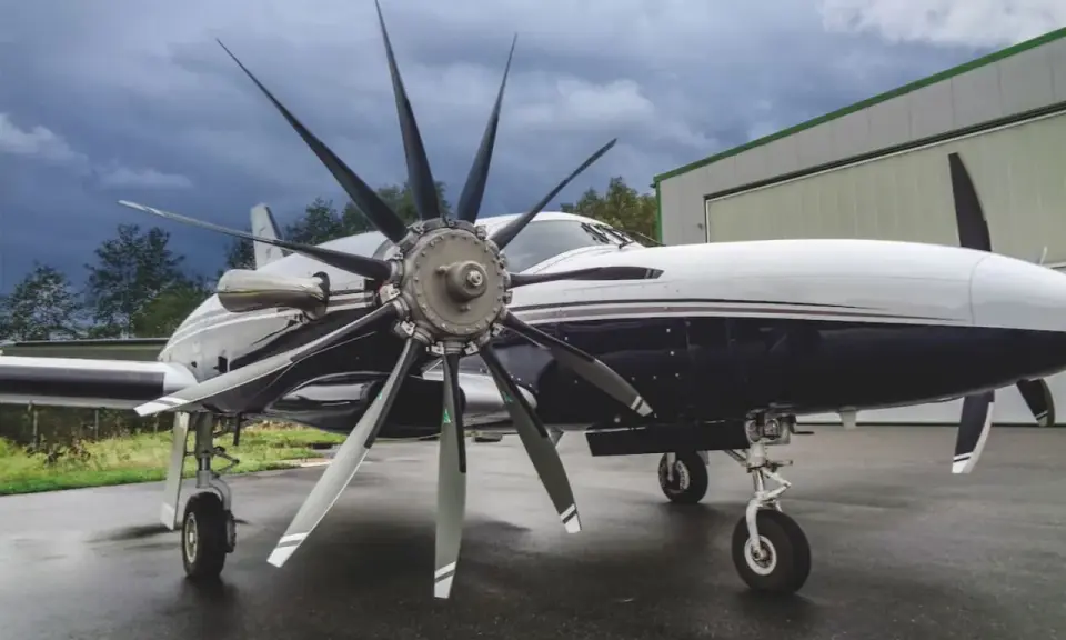 Ein elegantes Flugzeug mit einem beeindruckenden 4-Blatt-Propeller steht auf nassem Asphalt vor einer Halle.
