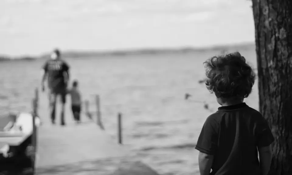 Bambino con capelli ricci guarda verso un molo sfocato dove camminano due figure. Un senso di isolamento, quasi un attaccamento disorganizzato, aleggia nell'aria.