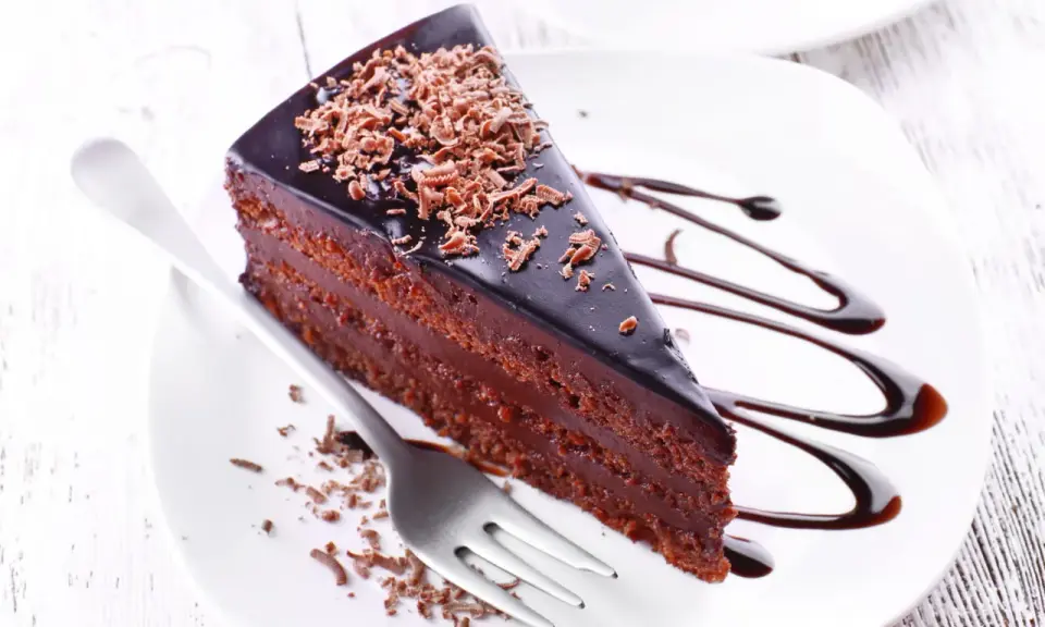 Une part de gâteau aux chocolats, nappée de glaçage brillant et garnie de copeaux de chocolat, est servie sur une assiette blanche avec un filet de sauce chocolat.