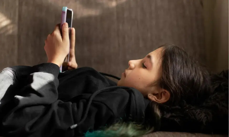 Ein Mädchen liegt auf einer Couch und schaut auf ihr Smartphone. Sie ist in ein schwarzes Kapuzensweatshirt gekleidet. Dies spiegelt aktuelle Trends der Jugend wider.