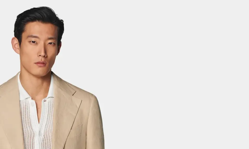A man models a tan linen blazer over a white textured polo shirt, perfect for a smart casual dress code for a wedding.