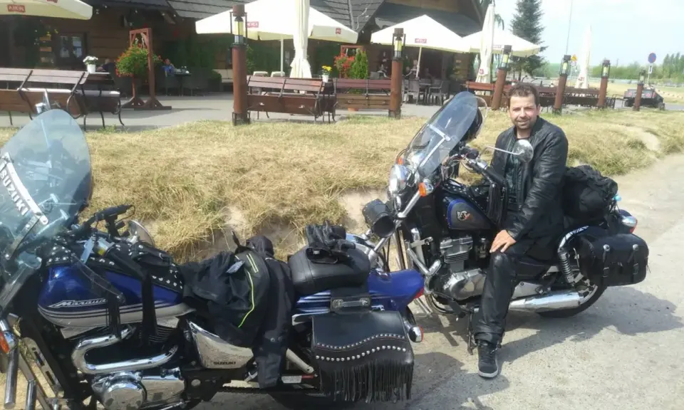 Motocyklista w skórzanej kurtce i spodniach odpoczywa przy swoim motocyklu. W tle karczma "Bida", idealne miejsce na postój podczas odkrywania **tras motocyklowych lubelskich**.