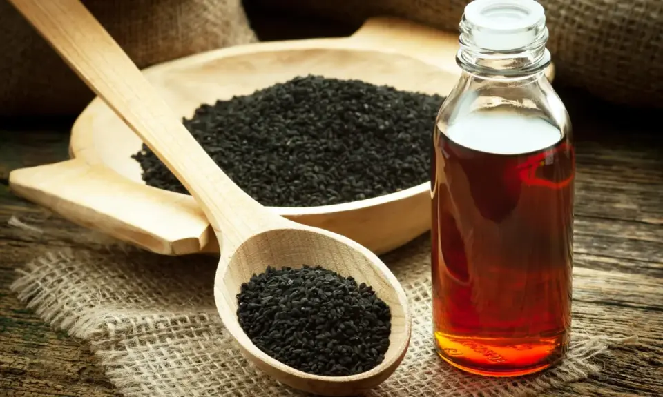 Huile de nigelle ambrée dans un flacon, à côté de graines noires dans une cuillère en bois. Découvrez les bienfaits de l'huile de nigelle.