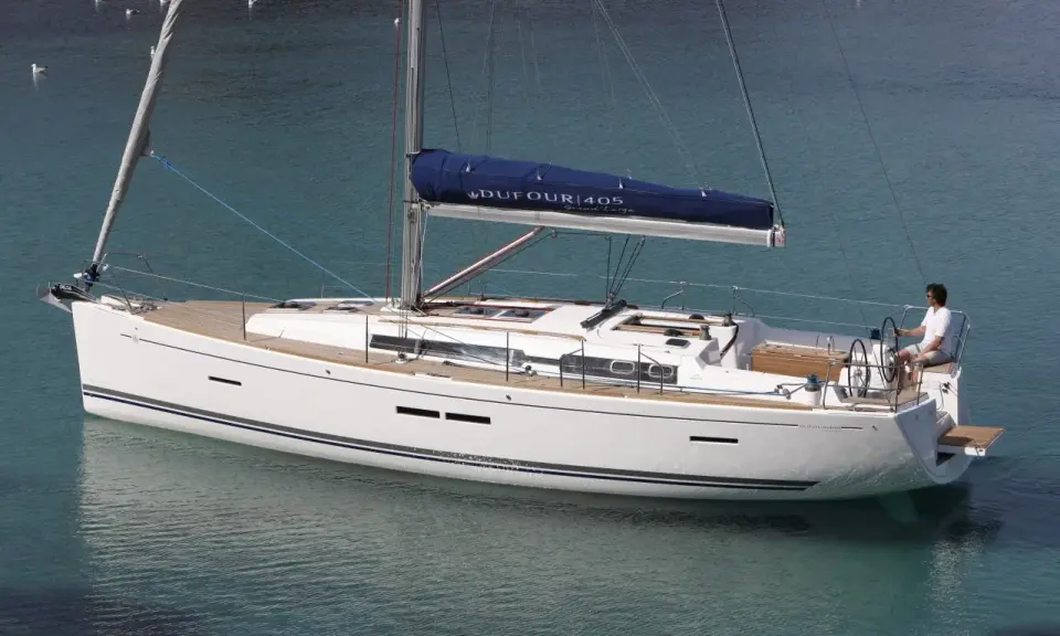 Un voilier blanc Dufour 405 Grand Large navigue sur une eau turquoise. Un homme est au poste de barre.