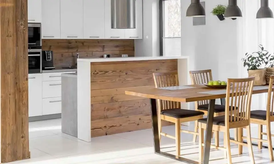 Cocina semiabierta al salón con barra de madera, encimeras blancas y electrodomésticos integrados. Mesa de comedor de madera con sillas.