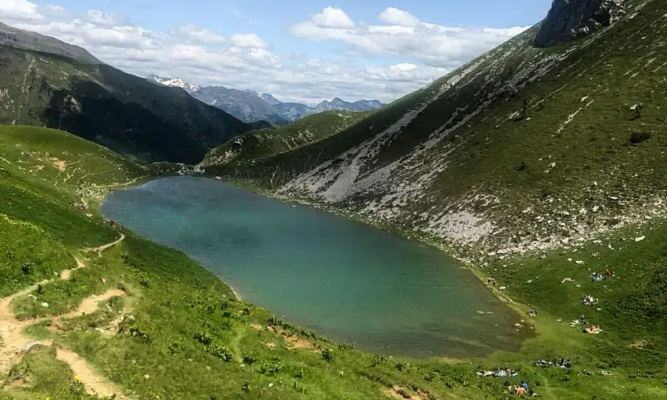 Un sentiero serpeggia tra prati verdi verso il lago Branchino, le cui acque turchesi riflettono le montagne circostanti. Alcune persone si godono un picnic sulle rive.