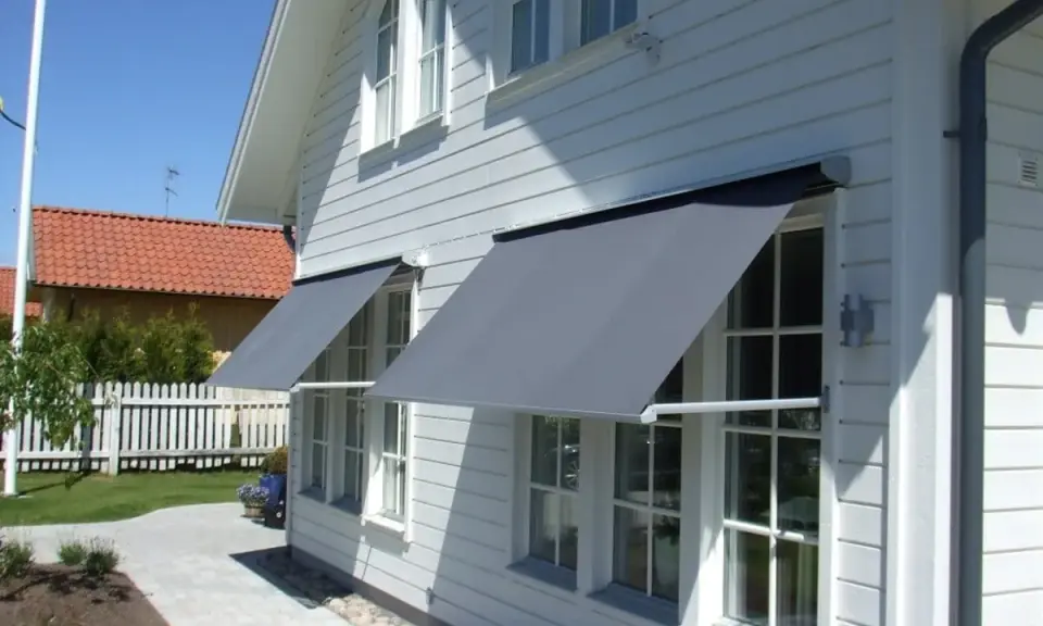 Grå markiser monterade på ett vitt trähus. Solen skiner och skapar skuggor på fasaden.