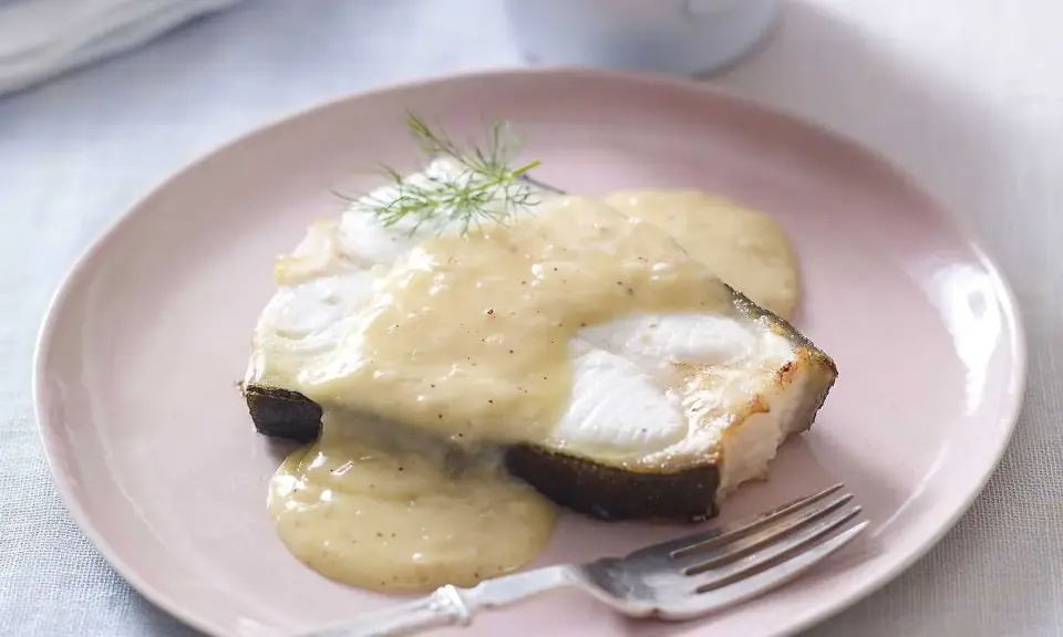 En bit stekt fisk med en krämig beurre blanc-sås och dill på en ljusrosa tallrik.