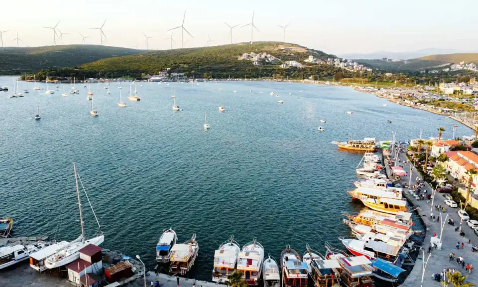 Port w Alacati, Turcja, pełen łodzi i żaglówek, z wiatrakami na wzgórzach w tle.