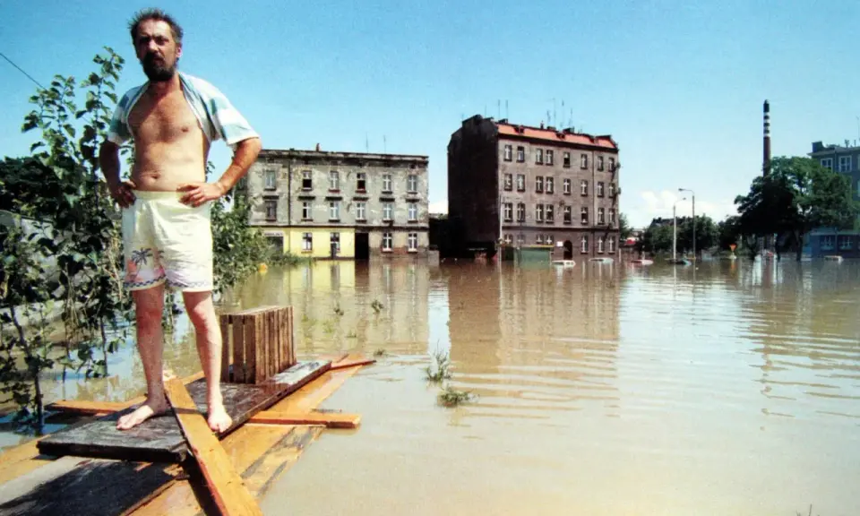 Mężczyzna na prowizorycznej tratwie w zalanej wodą ulicy. Budynki i samochody toną w brązowej wodzie. To obraz powodzi 1997.