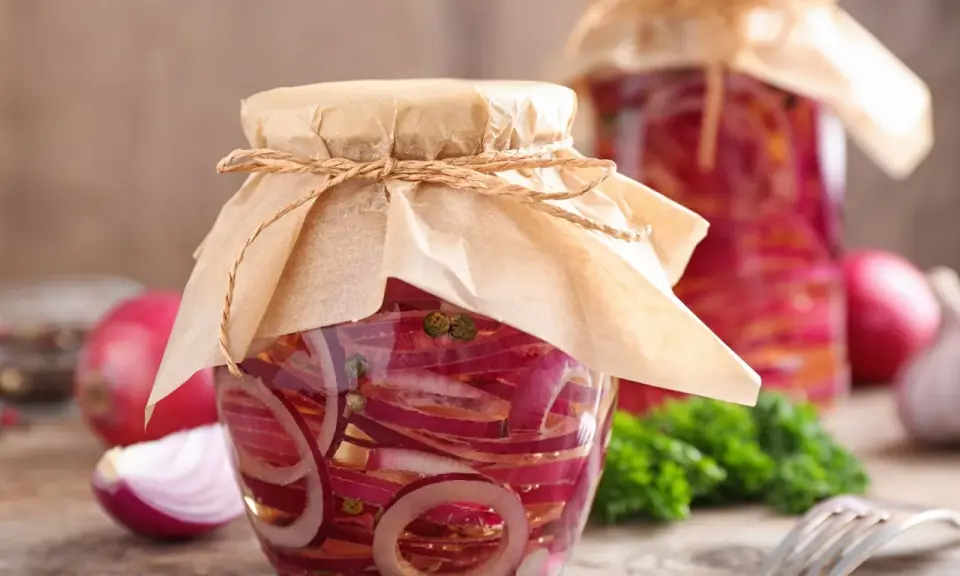 Zwei Gläser mit eingelegten Zwiebeln, verziert mit Papier und Schnur. Daneben liegen frische Zwiebeln und ein Gabel.