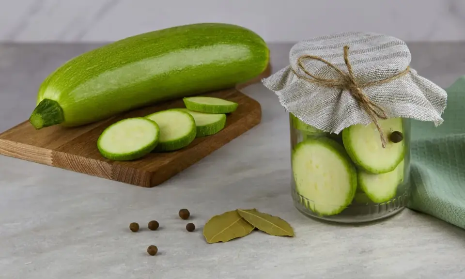 Frische Zucchini und Scheiben auf einem Holzbrett, daneben ein Glas mit eingelegten Zucchinischeiben. Perfekt zum Zucchini fermentieren.
