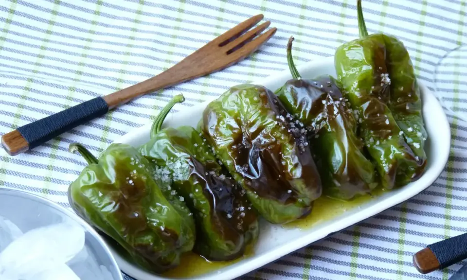 Délicieux **pieds de poivrons** verts, grillés et saupoudrés de gros sel, servis dans une assiette blanche avec une fourchette en bois.