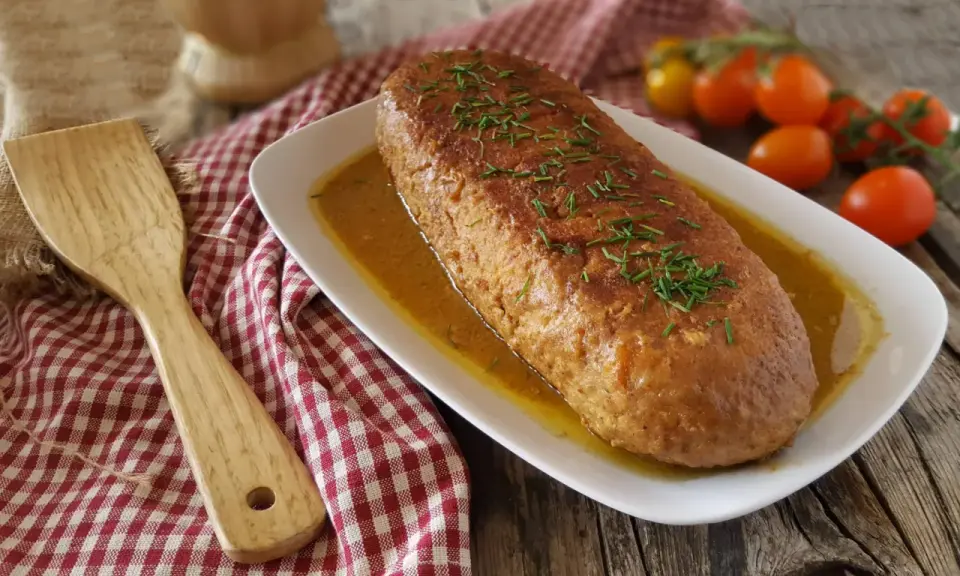 Polpettone in padella dorato e succulento, guarnito con erba cipollina, pronto per essere servito. Accanto, una spatola in legno e pomodorini freschi.