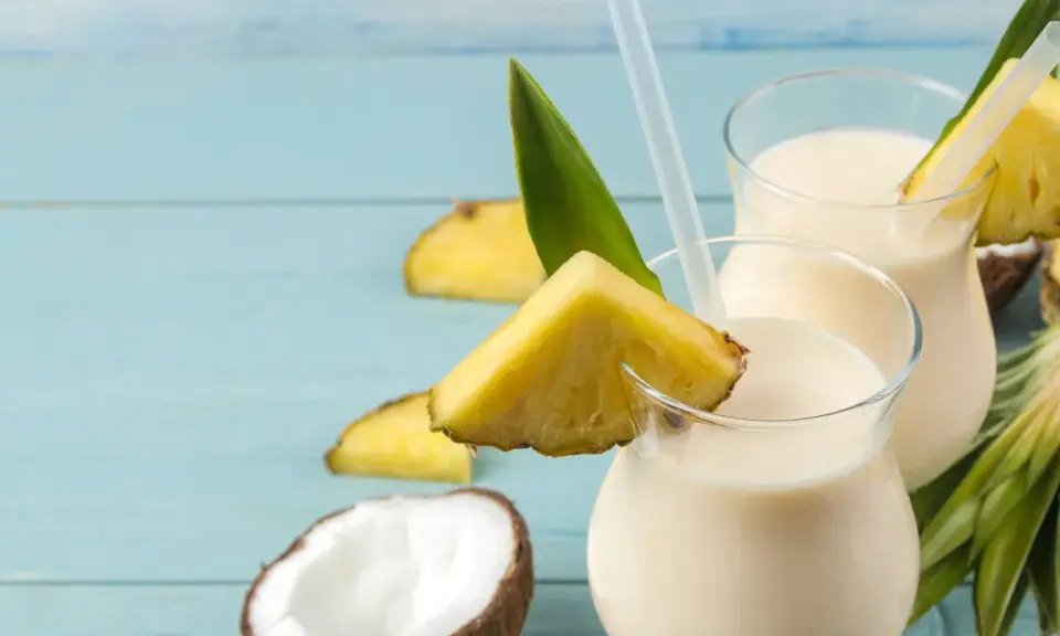 Dwa orzeźwiające koktajle Pina Colada, ozdobione kawałkami ananasa i liśćmi, na tle niebieskich desek. Idealny przepis na lato!
