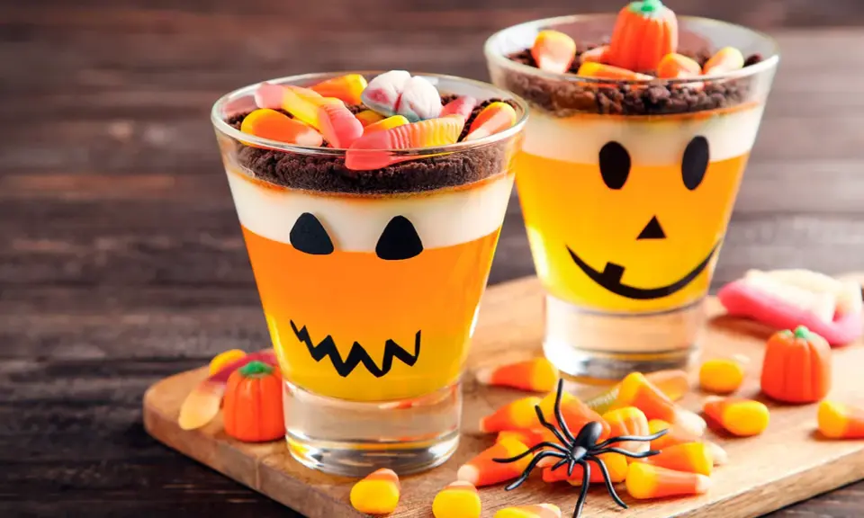 Deliciosa gelatina Halloween para niños, con capas de naranja y blanco, decorada como calabaza y cubierta de dulces.