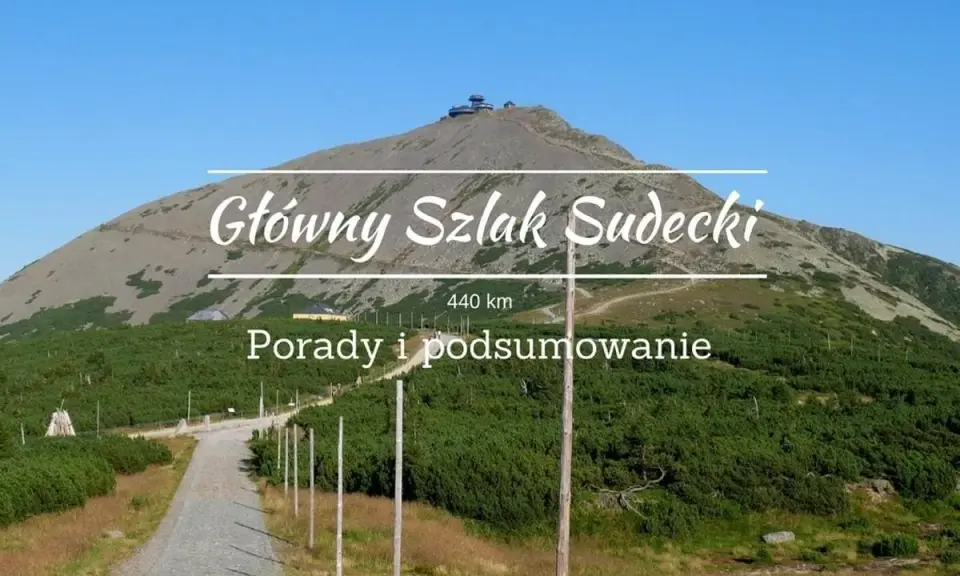 Kamienista ścieżka prowadzi na szczyt góry, gdzie widać budynek. Nad krajobrazem widnieje napis "Główny Szlak Sudecki".