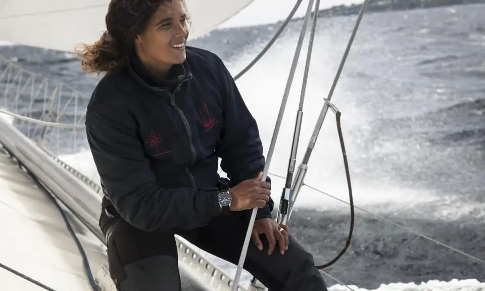 Marie Tabarly, souriante, tient la barre d'un voilier, le vent dans ses cheveux, naviguant sur une mer agitée.