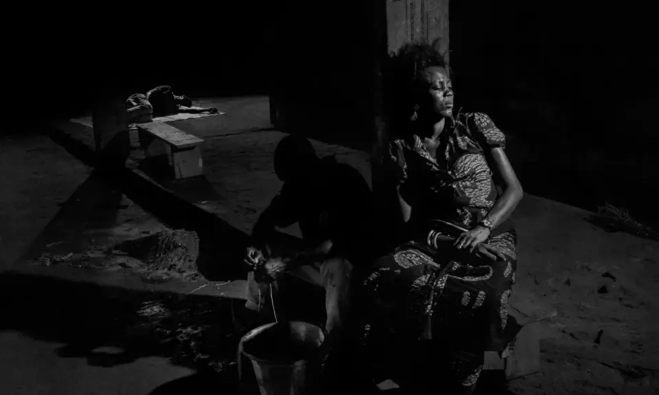 A woman in a patterned dress sits in shadow, eyes closed, while a man works with a bucket in the foreground. Alex Majoli captures a moment of quiet resilience.
