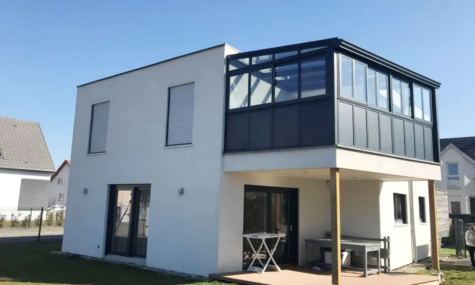Maison moderne avec une veranda 2 niveaux vitrée, offrant une extension lumineuse. Terrasse en bois en contrebas.