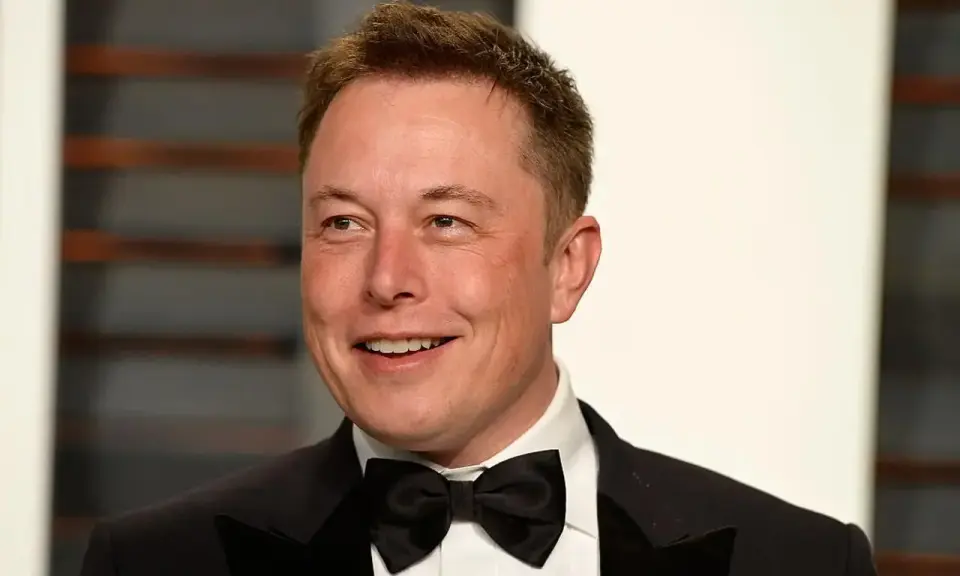 Uśmiechnięty Elon Musk w smokingu z muszką. Wiek nie jest widoczny, ale jego twarz wyraża pewność siebie.