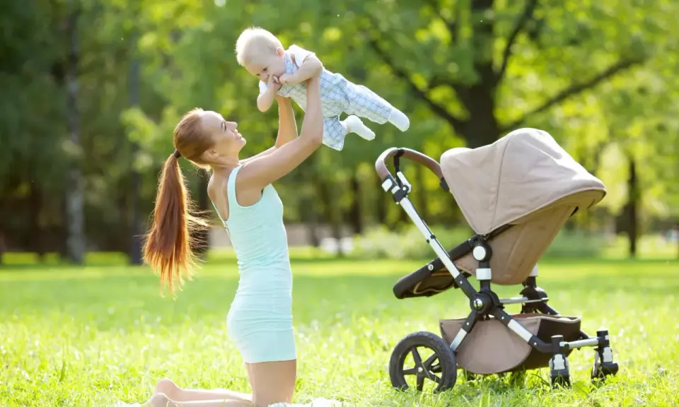Uśmiechnięta mama podrzuca dziecko w powietrze na trawie obok wózka.