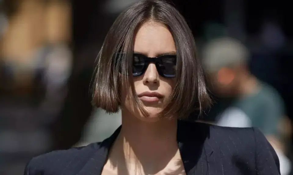 Une femme au regard intense arbore une coupe carré droit dégradé, accentuée par des lunettes de soleil noires et un blazer chic.
