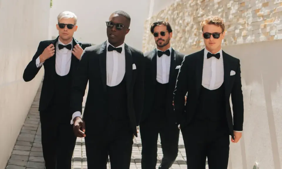 Four men stride confidently in a modern black tie dress code, sporting sharp tuxedos and sunglasses.