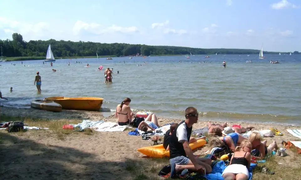 Ludzie odpoczywają na plaży i kąpią się w jeziorze. W tle żaglówki i las. To typowe boszkowo atrakcje w letni dzień.