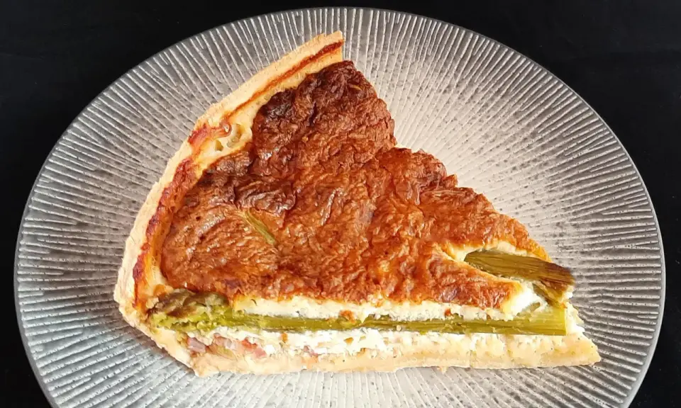 Tranche de tarte aux asperges blanches, garnie d'une préparation crémeuse et dorée, sur une assiette texturée.