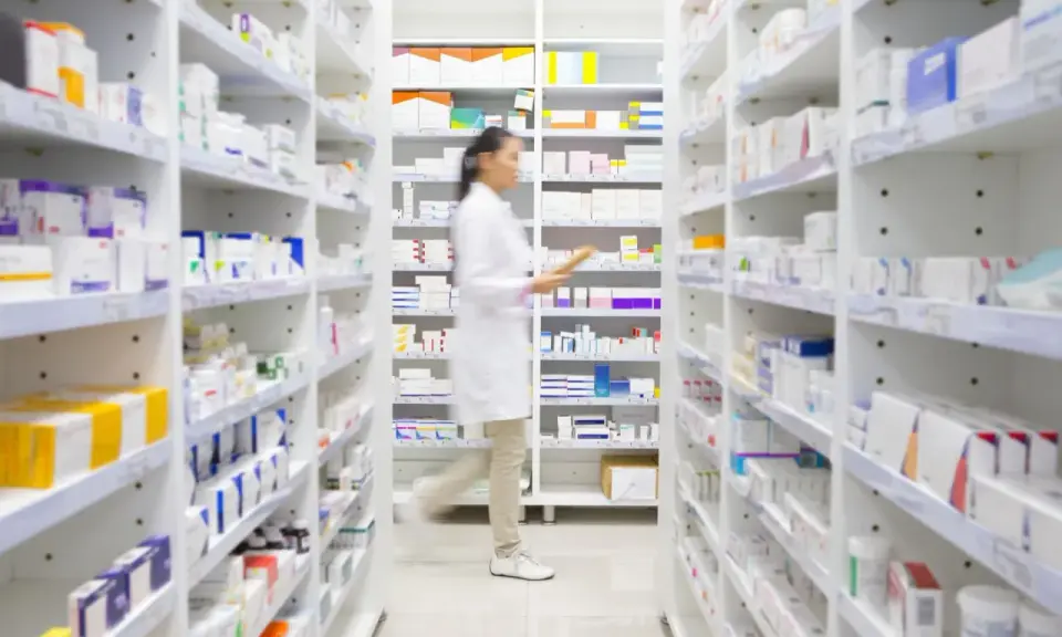 Farmaceutka w białym fartuchu idzie między regałami pełnymi leków. Widać, na co **emanera na co** się przygotowuje, zapewne do wydania pacjentowi.