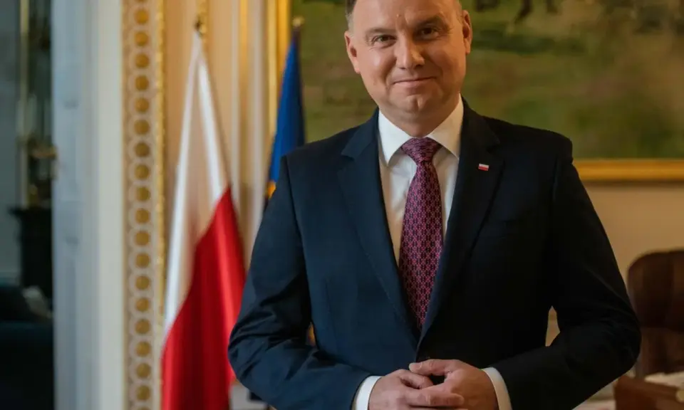 Prezydent Andrzej Duda w garniturze, z polską flagą w tle, czeka na moment, kiedy prezydent podpisze emerytury stażowe.
