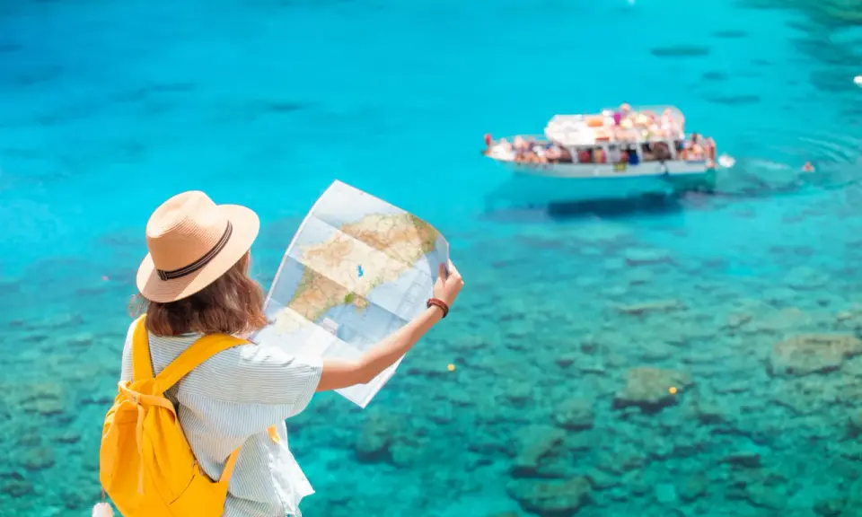 Ragazza con zaino giallo e cappello di paglia studia una mappa, pensando a quanti soldi portare in crociera, con mare turchese e barca sullo sfondo.