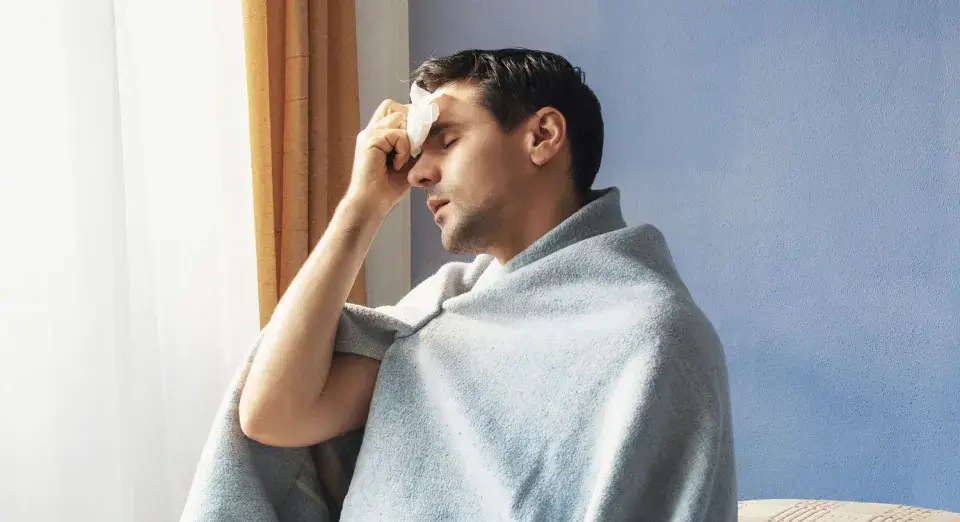 Uomo con panno umido sulla fronte, avvolto in una coperta, che sembra soffrire di sudori freddi.