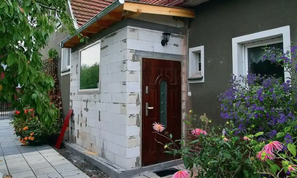 Budowa dobudowy wiatrołapu z bloczków, z nowymi oknami i drzwiami, otoczona zielenią.