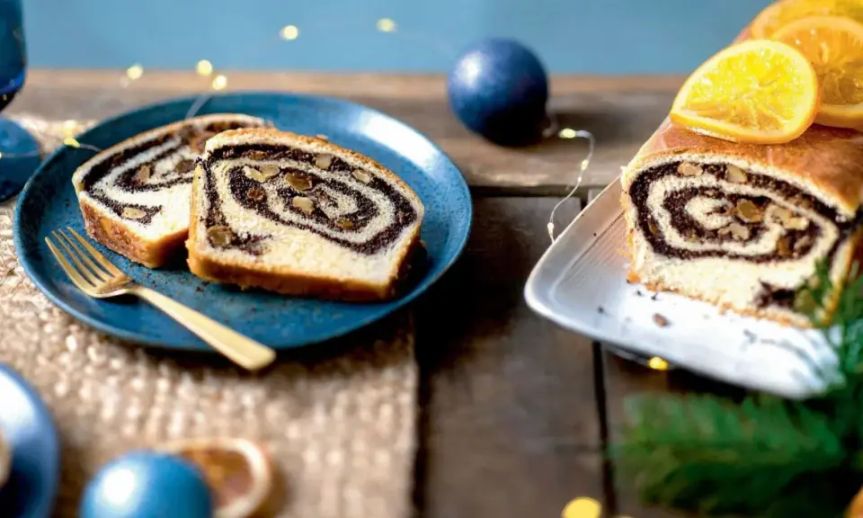 Tranches de roulé au pavot, garni de noix, sur une assiette bleue avec une fourchette dorée. Un autre roulé décoré d'orange est à droite.