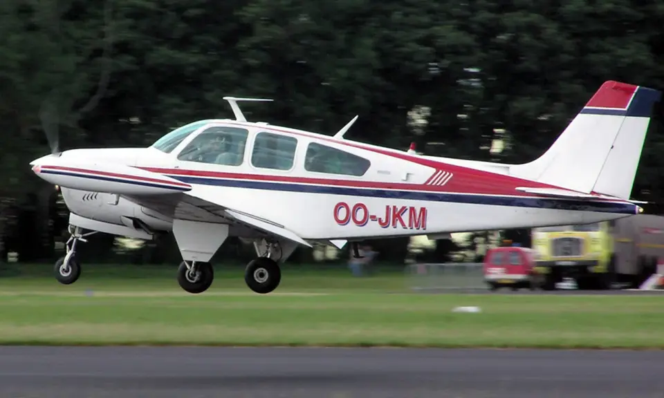 Eine weiße Beechcraft Bonanza mit roten und blauen Streifen hebt vom Gras ab.
